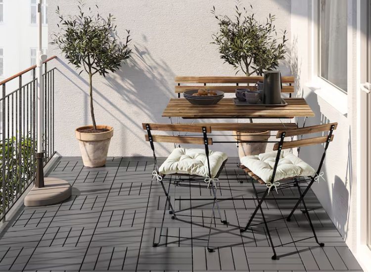 Wooden foldable bench with seating pillows and chairs on a balcony, surrounded by two small plants for a cozy atmosphere.